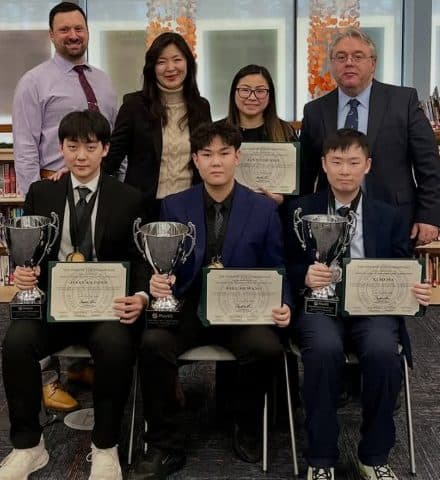 Three students in suits sit in a row, each holding a silver cup trophy and a printed certificate, with four adults standing behind them in front of library bookshelves.
