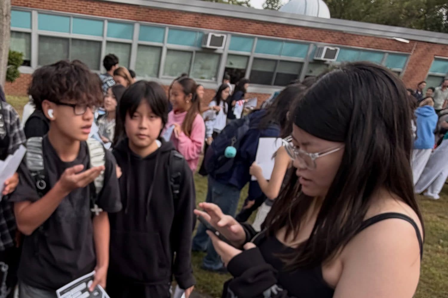 Students standing close together on the grass beside the club’s table, holding printed flyers and phones and talking, with a low brick school building behind them.