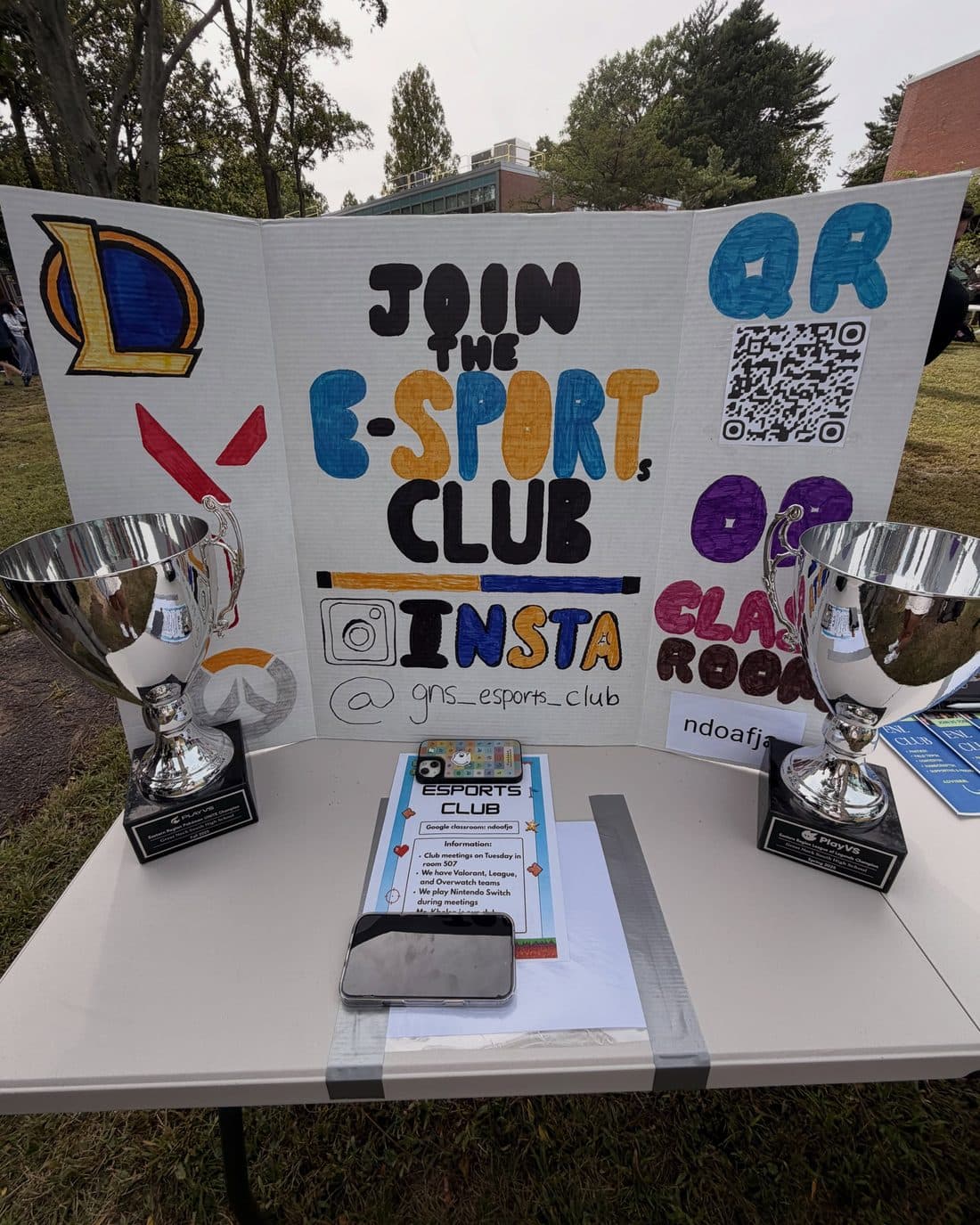 A hand-lettered display board reading “Join the E-Sport Club” at an outdoor club fair, with drawn game logos and a QR code on it, and a tall silver cup trophy standing on the folding table at either side.